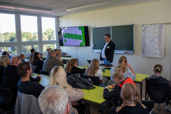 Der Schulleiter stellt in einem gefüllten Klassenraum das Schulkonzept vor